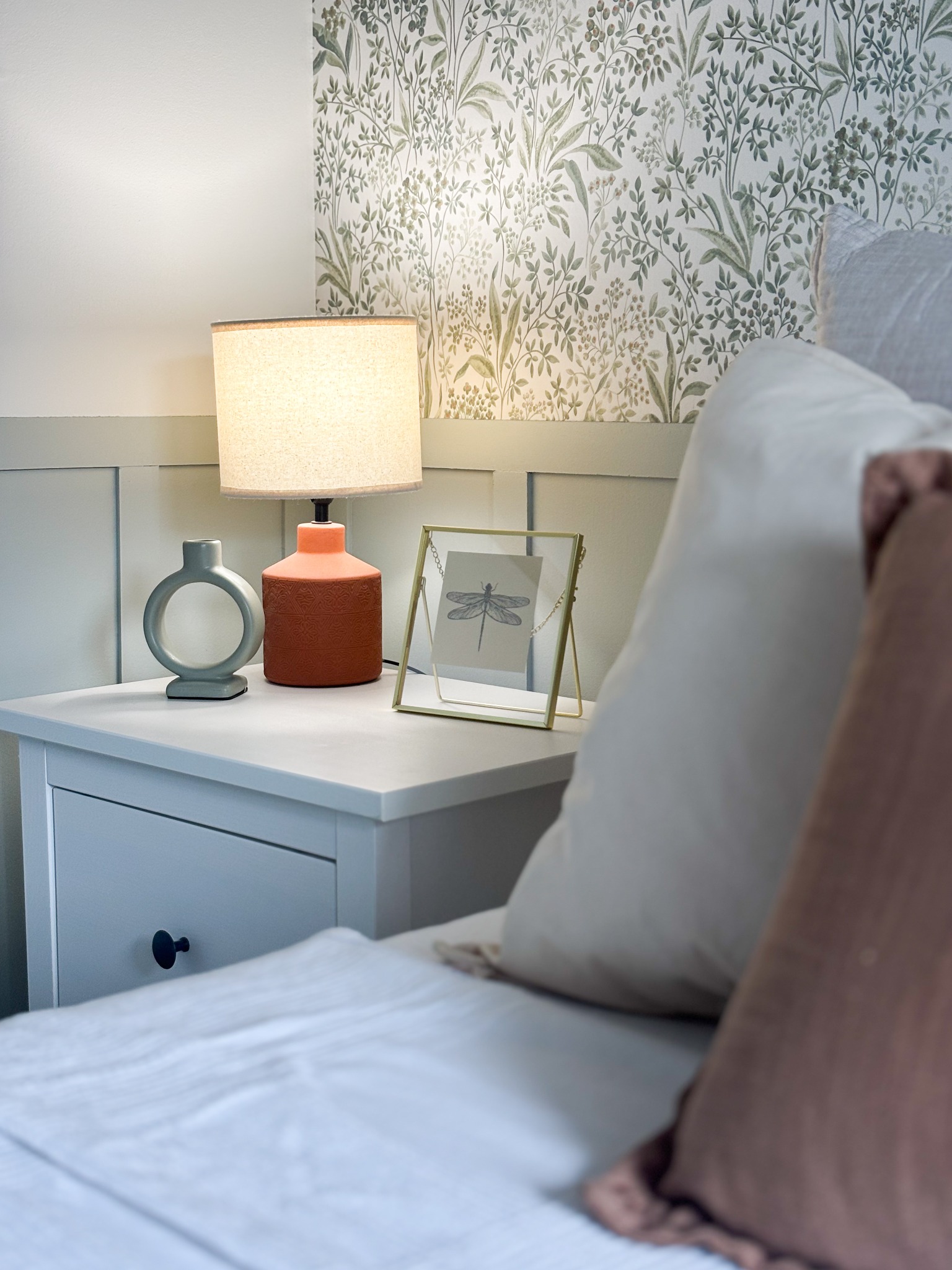 Szafka nocna z lampką, ceramiczną wazą w kształcie okręgu i ramką ze szkła, na tle ściany z tapetą w roślinny wzór, uzupełniona poduszkami na łóżku.