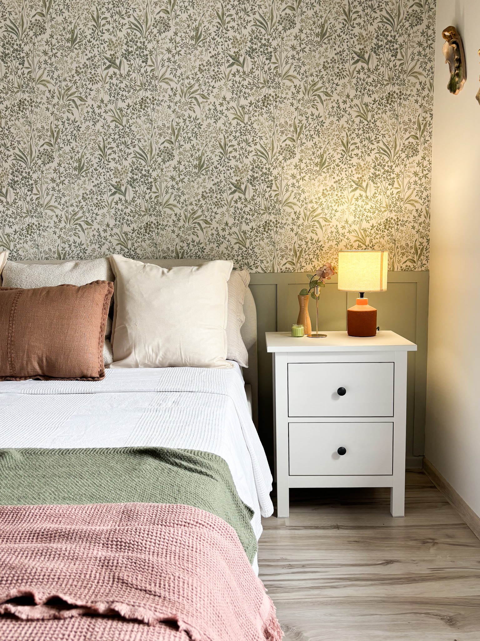Sypialnia z łóżkiem, stolikiem nocnym z lampką i tapetą w roślinny wzór, utrzymana w stonowanej kolorystyce z akcentami brązu i różu.