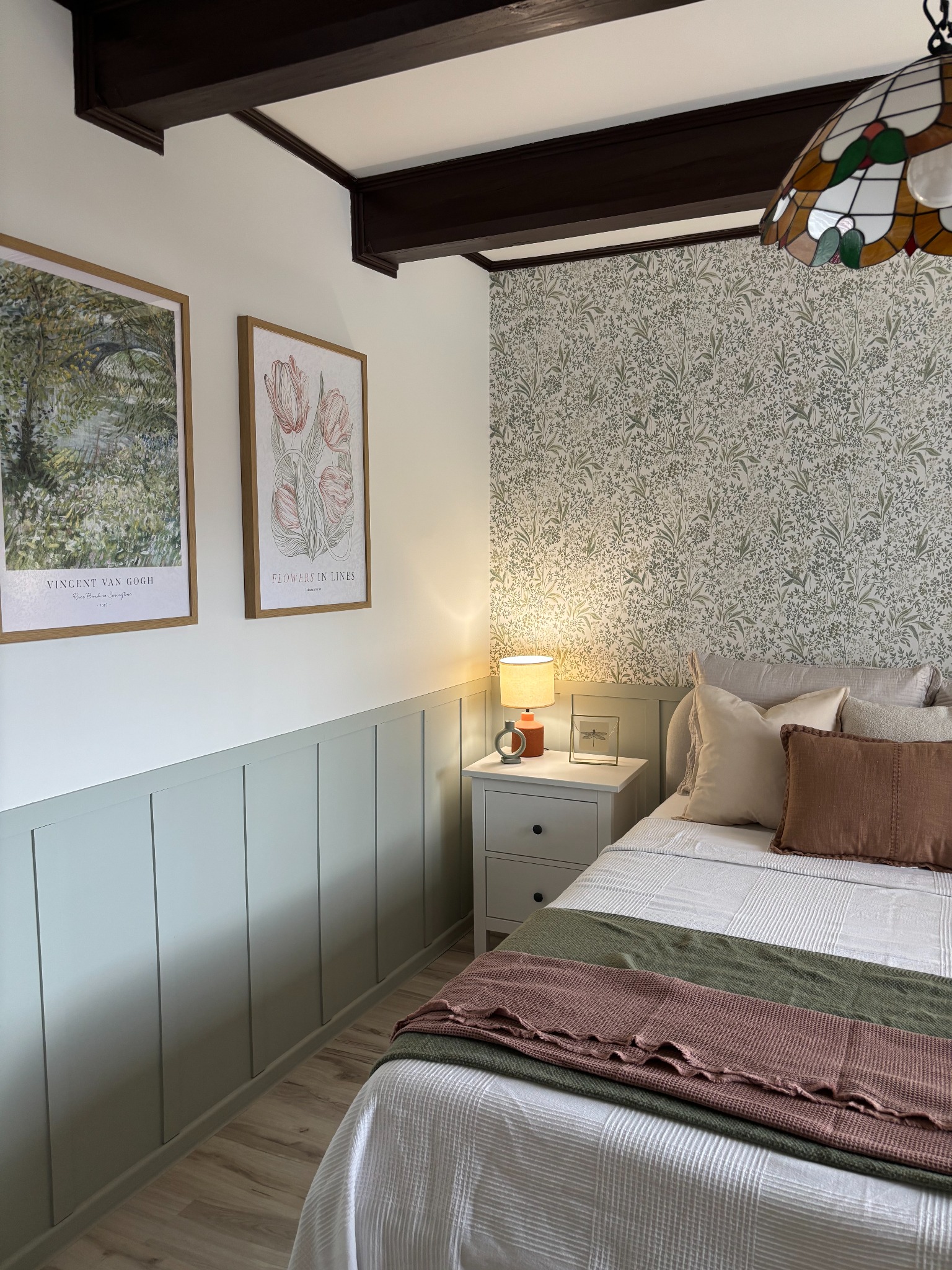 Przytulna sypialnia w stylu cottagecore z akcentami vintage: wzorzysta tapeta, drewniane belki, obrazy w ramach, lampka nocna z pomarańczową podstawą i patchworkowa narzuta na łóżku.