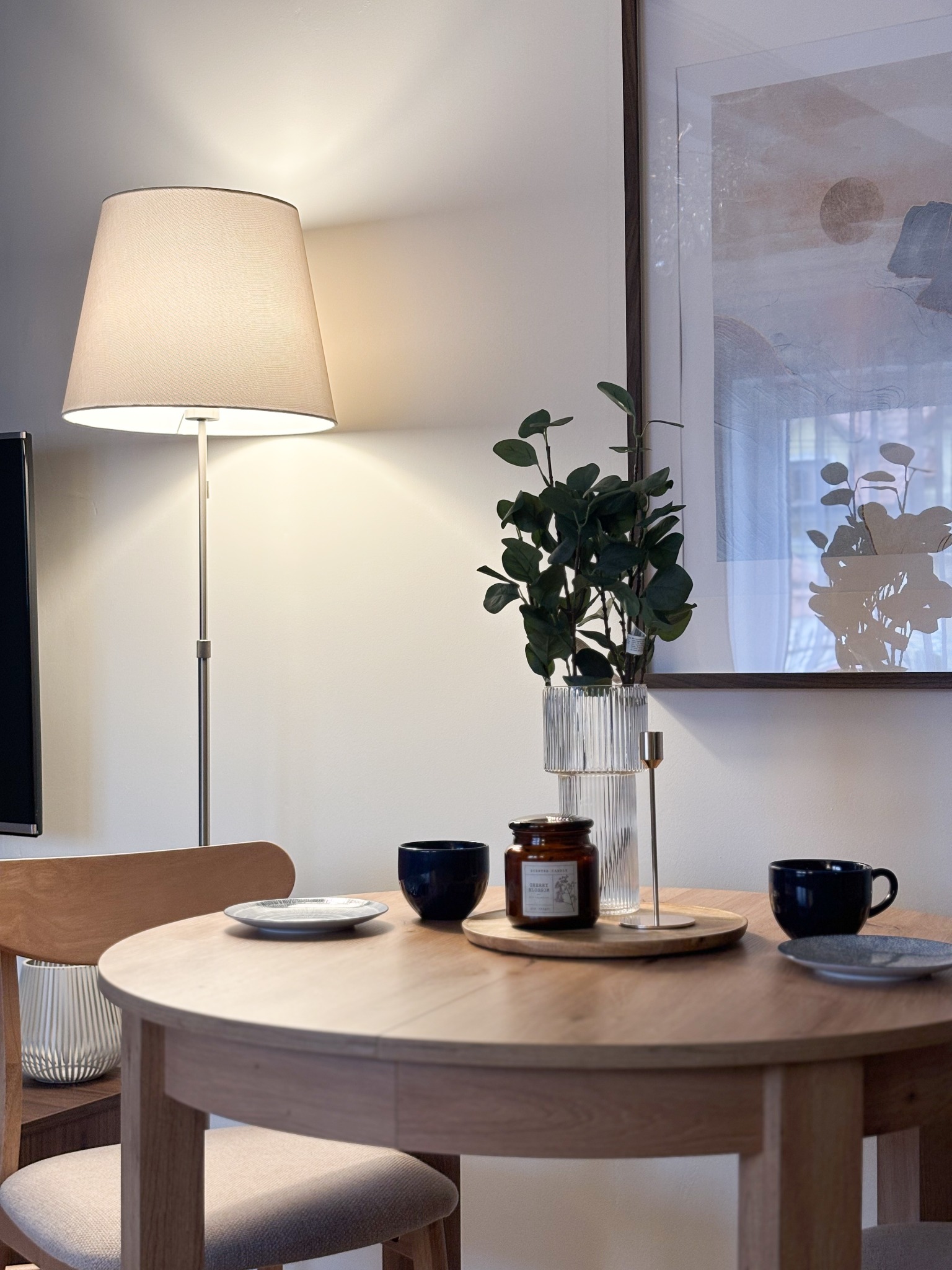 Okrągły drewniany stół z nakryciem: talerze, filiżanki, świeca zapachowa i wazon z zielonymi gałązkami, oświetlony lampą podłogową, w tle abstrakcyjny obraz w ramie.