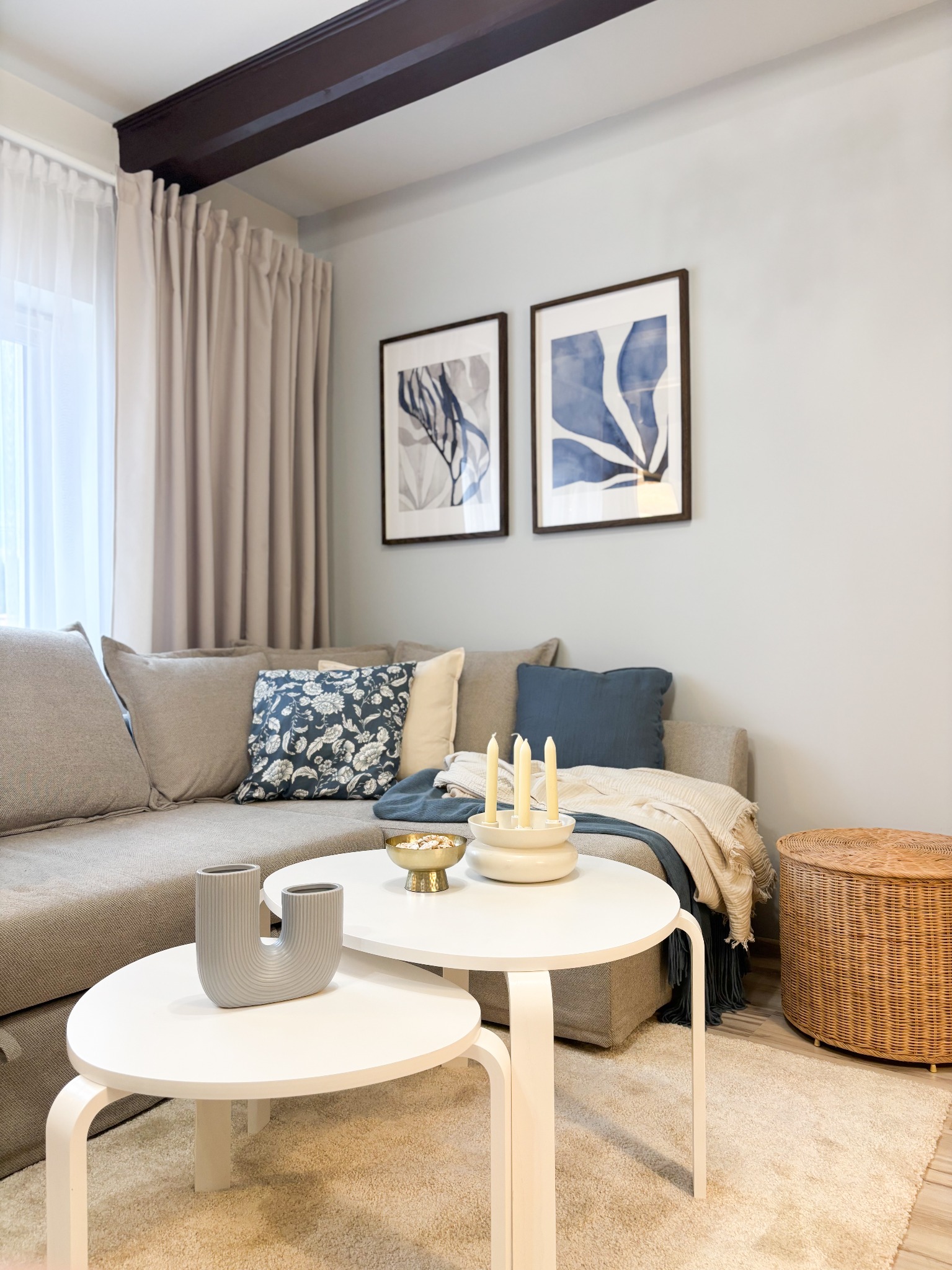 Jasny salon z narożną sofą, dwoma okrągłymi stolikami, dekoracjami i dwoma obrazami na ścianie, podkreślony ciemną belką stropową.