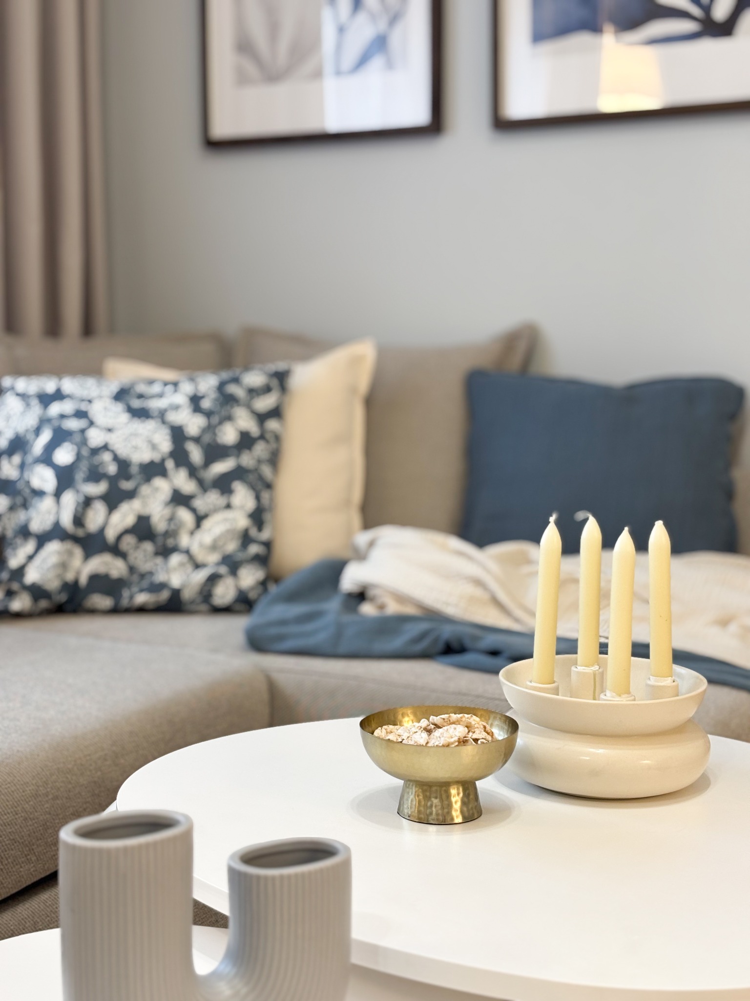 Neutralna aranżacja salonu z beżową sofą, poduszkami w odcieniach błękitu i beżu, białym stolikiem kawowym z dekoracjami: złotą miseczką z potpourri i ceramicznym świecznikiem z czterema świecami.