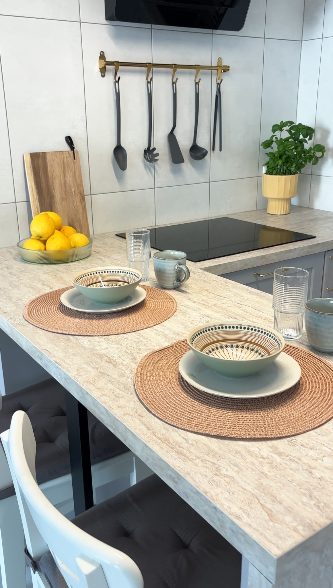 Ustawiony stół z dwoma nakryciami, miseczki w odcieniach beżu i zieleni, szklanki z prążkowaną fakturą, ceramiczne kubki, owoce w szklanej misie, czarne przybory kuchenne wiszące na złotej listwie...