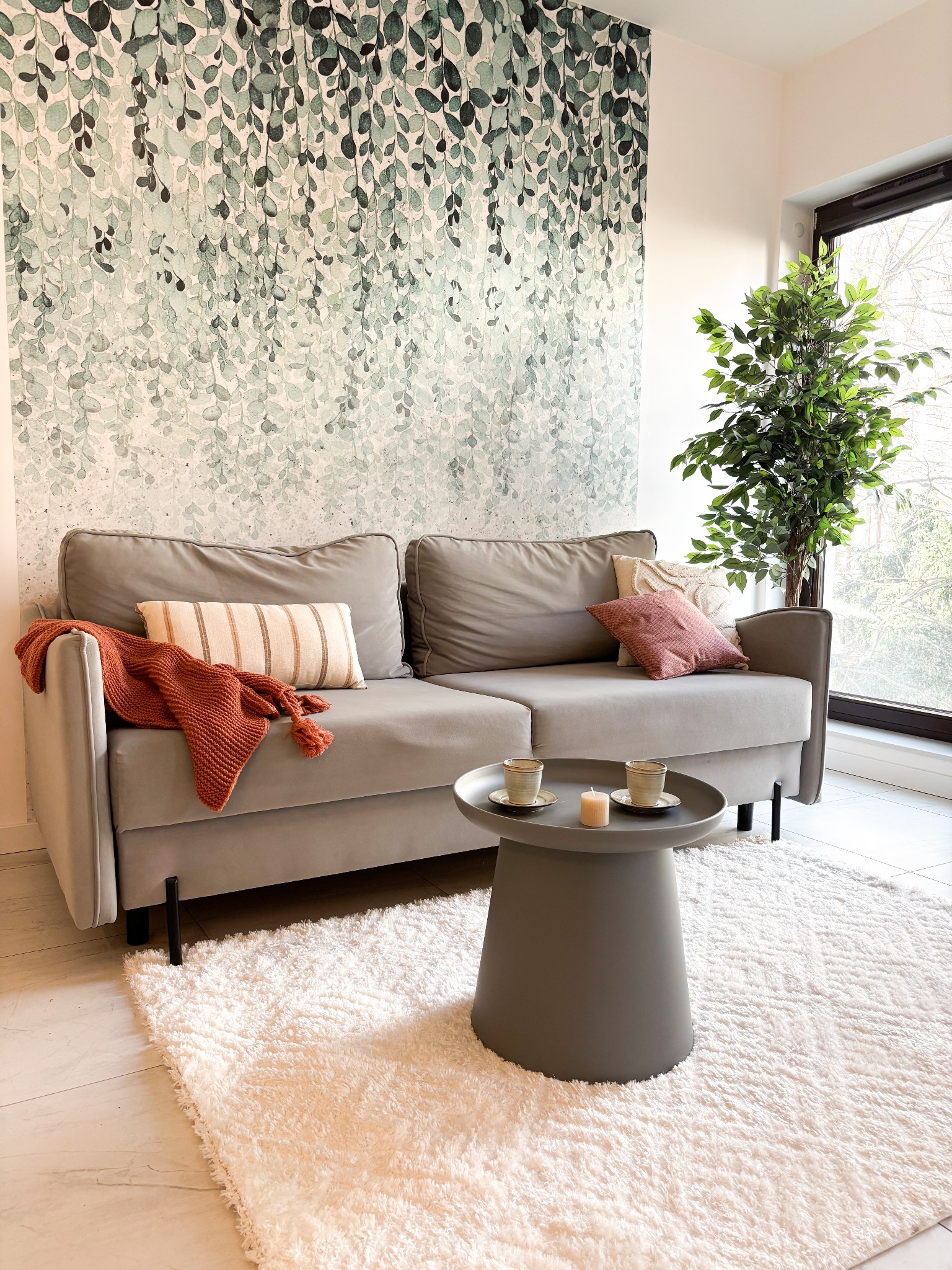 Jasnoszara sofa z poduszkami i pomarańczowym pledem, okrągły stolik kawowy z dwoma filiżankami i świeczką na puszystym dywanie, na ścianie tapeta z motywem liści, obok okna stoi zielona roślina.