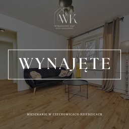 PRETTY HOME Anna Sztorc - Salon z czarną sofą, okrągłym stolikiem kawowym i drewnianą podłogą, oświetlony lampą stojącą, z napisem 'Wynajęte' na zdjęciu, logo agencji nieruchomości w górnej części kadru.