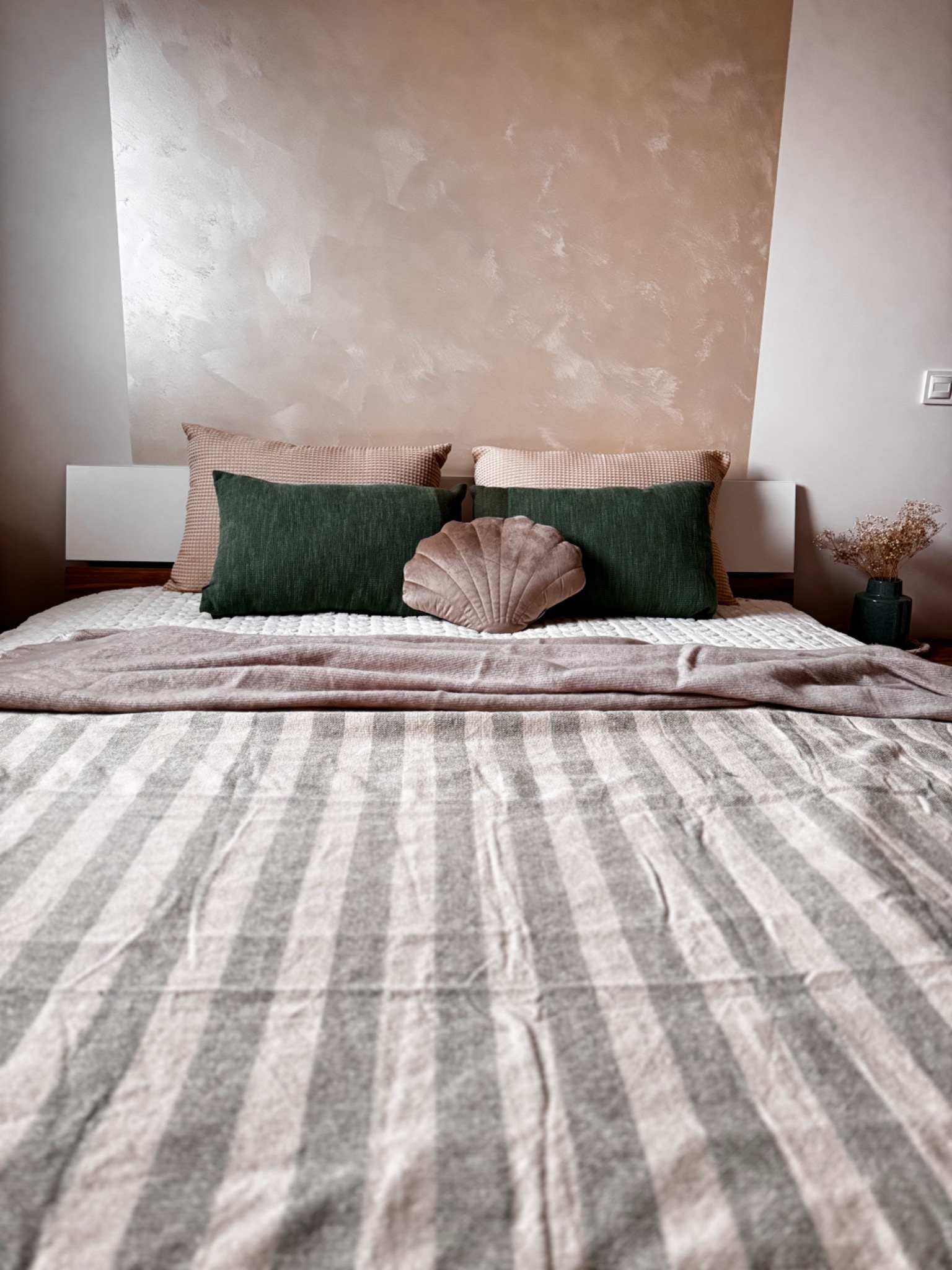Sypialnia z łóżkiem w paski, zielonymi poduszkami, dekoracyjną poduszką w kształcie muszli i wazonem z suszonymi kwiatami na tle ściany w odcieniach beżu i szarości.