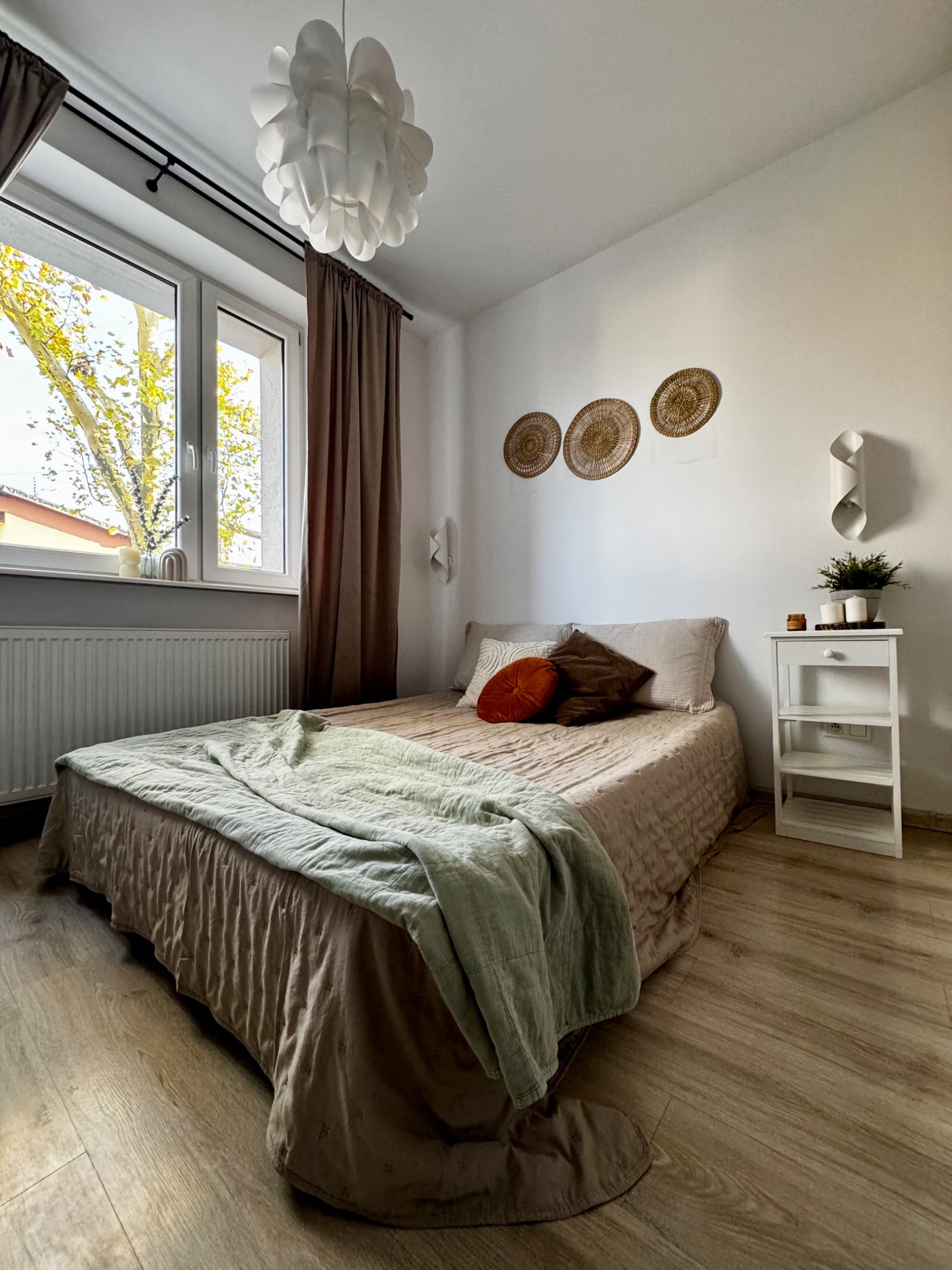 Przytulna sypialnia z łóżkiem w beżowej pościeli, zielonym pledem i dekoracyjnymi poduszkami, trzy plecione dekoracje ścienne nad łóżkiem, biała szafka nocna z rośliną i świecami, okno z widokiem...