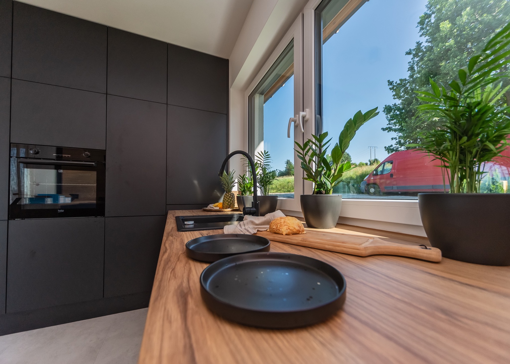 Nowoczesna kuchnia z czarnymi matowymi szafkami, drewnianym blatem, czarnym zlewem i armaturą, z roślinami w doniczkach na parapecie okiennym, na którym leżą również talerze i ananas