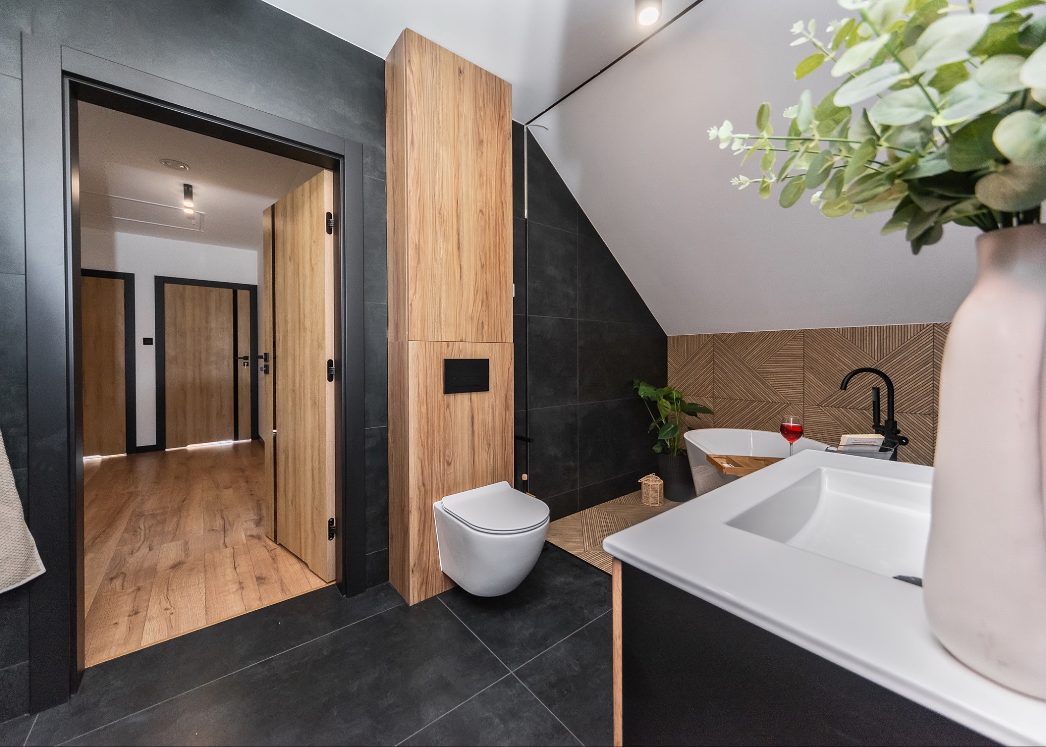 Nowoczesna łazienka w Czechowicach-Dziedzicach z czarnymi płytkami, drewnianymi akcentami, wiszącą toaletą i otwartymi drzwiami ukazującymi parkiet w dalszej części mieszkania.