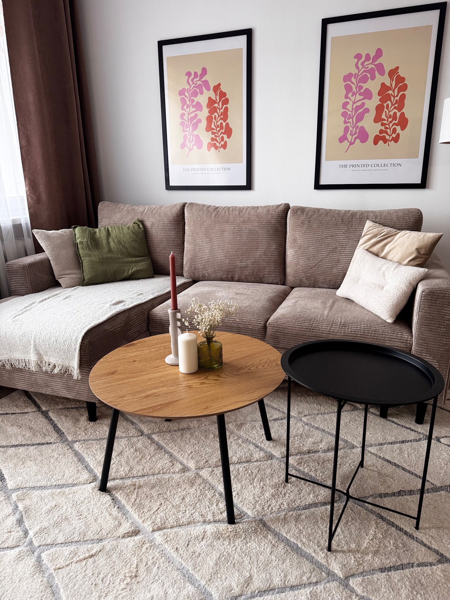 Beżowa sofa z poduszkami, okrągły drewniany stolik kawowy z dekoracjami i czarny stolik pomocniczy na wzorzystym dywanie w salonie z plakatami na ścianie.
