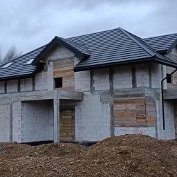 Skra bud - Surowy stan domu jednorodzinnego z szarej kostki, świeżo pokrytego czarną dachówką i orynnowaniem, z deskami zasłaniającymi otwory okienne i drzwiowe, na tle pochmurnego nieba.