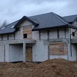 Surowy stan domu jednorodzinnego z szarej kostki, świeżo pokrytego czarną dachówką i orynnowaniem, z deskami zasłaniającymi otwory okienne i drzwiowe, na tle pochmurnego nieba.