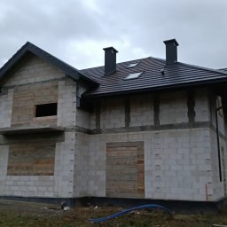 Skra bud - Dom w trakcie budowy, z szarą dachówką, widoczne kominy i okna dachowe, ściany z betonu komórkowego z zabezpieczonymi otworami okiennymi i drzwiowymi deskami.