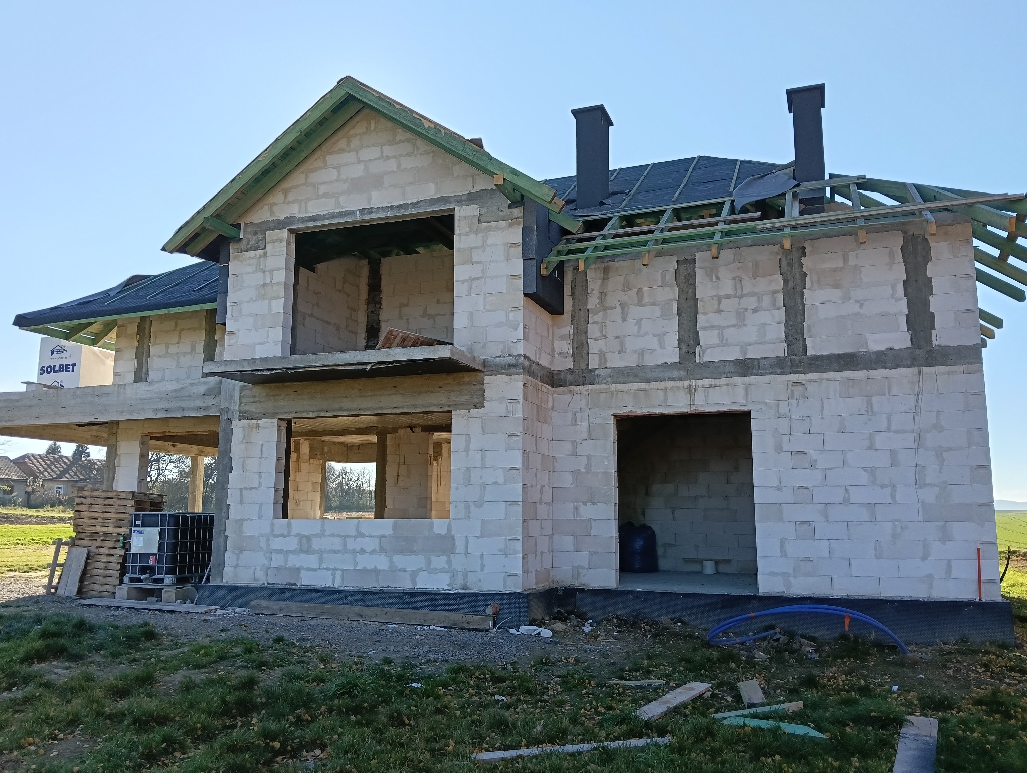 Niedokończony dwupiętrowy dom z białego pustaka z częściowo położonym czarnym pokryciem dachowym i widoczną zieloną więźbą, czarne kominy, palety i zbiornik na wodę obok budynku.