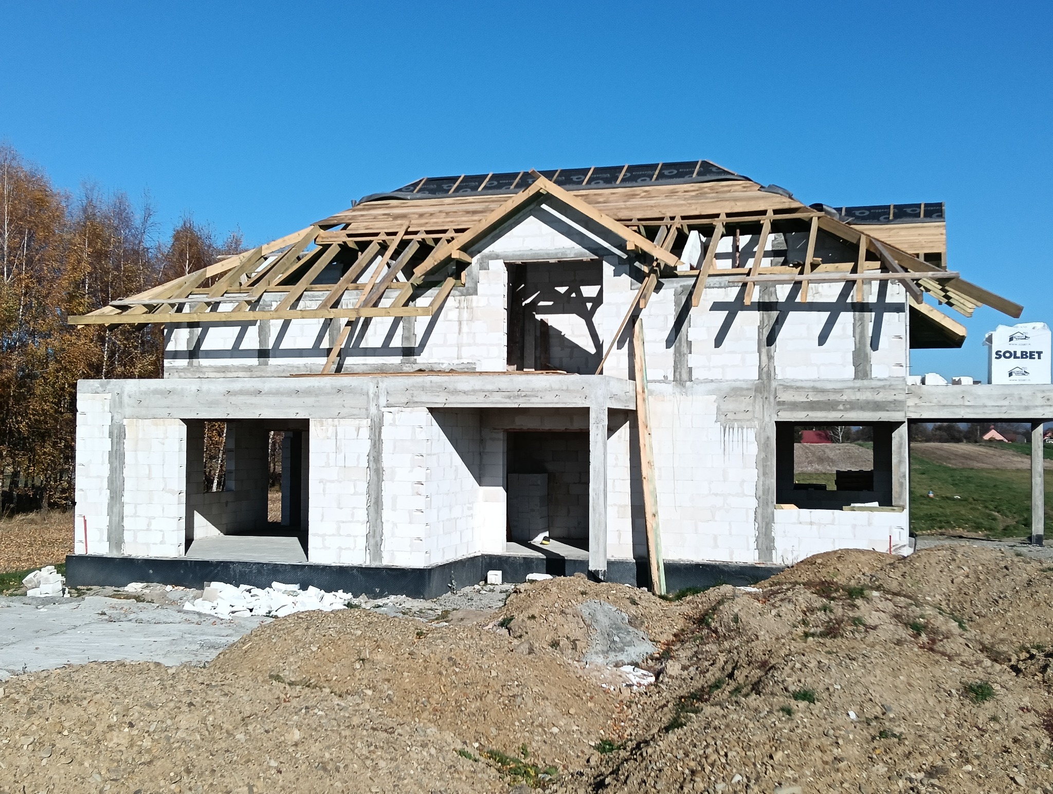 Wznoszony dom z białych bloczków, widoczna drewniana więźba dachowa i czarna membrana dachowa, w tle drzewa bez liści i błękitne niebo.