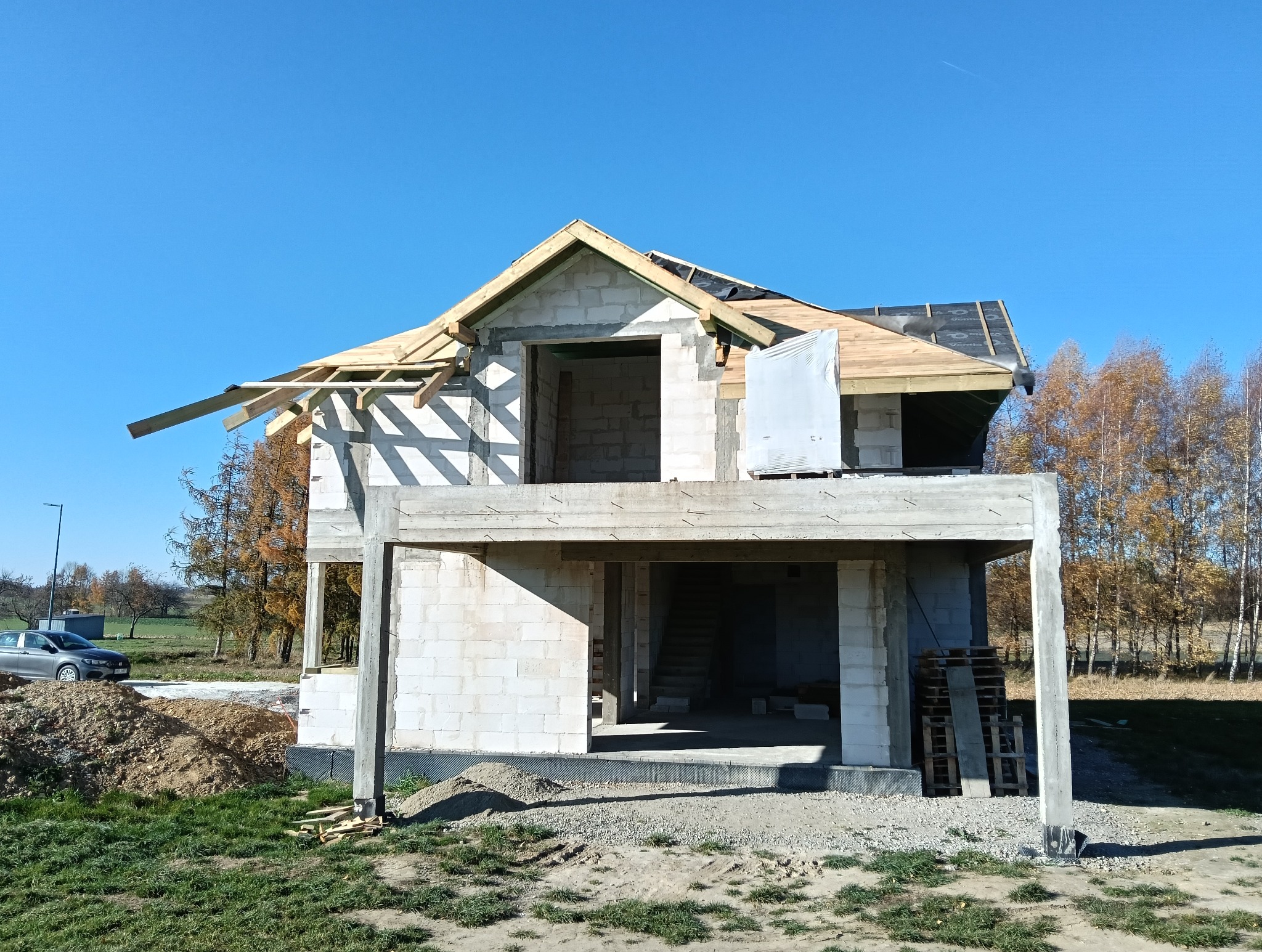 Dom w trakcie budowy, widoczna surowa konstrukcja dachu z drewnianymi belkami, częściowo pokryta membraną dachową, ściany z białego bloczka betonowego, otoczenie: trawa, drzewa i niebieskie niebo.