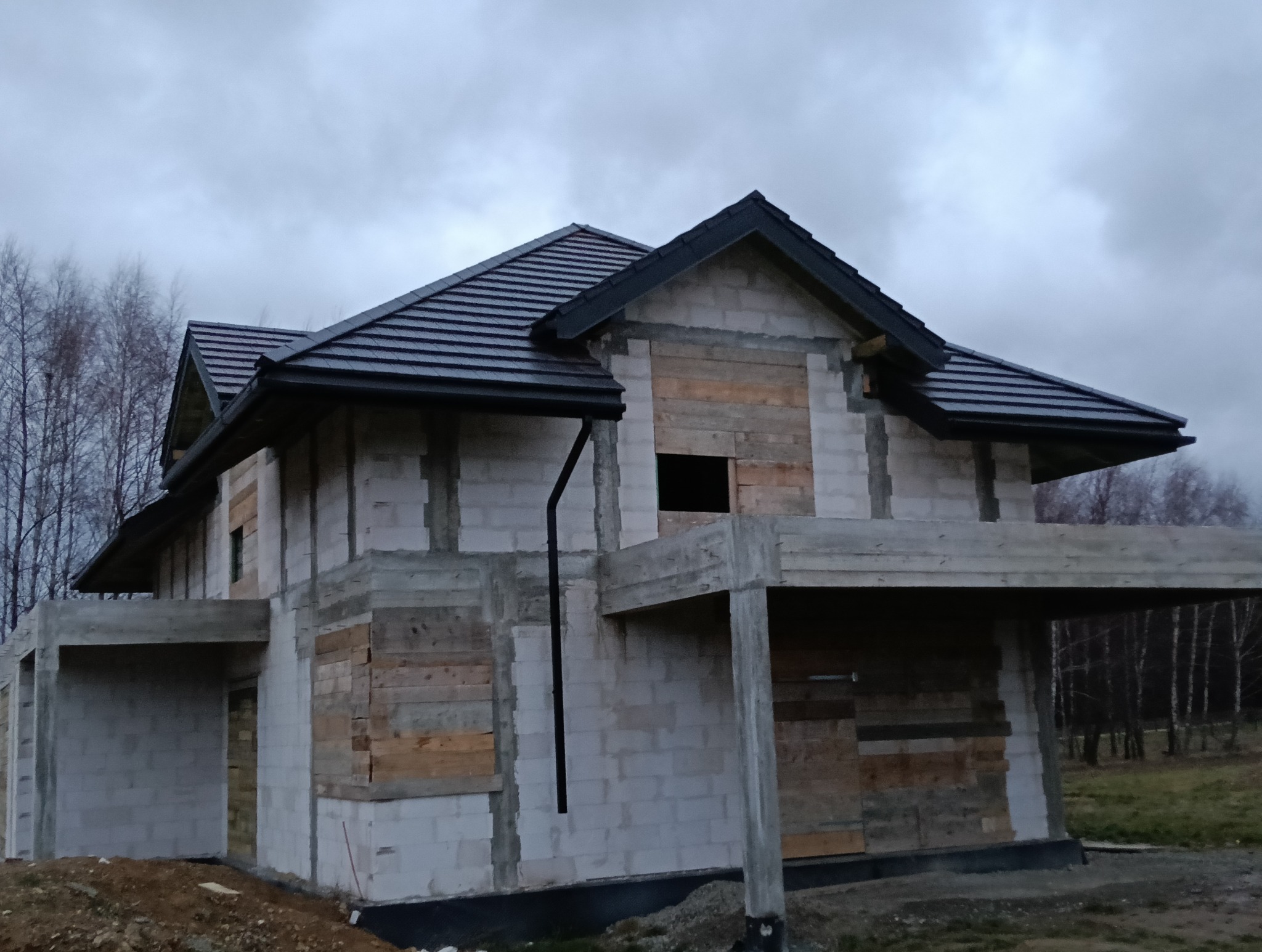 Surowy stan domu w trakcie budowy z ciemną dachówką, widoczne rynny i zabezpieczone okna deskami, szara pogoda w tle.
