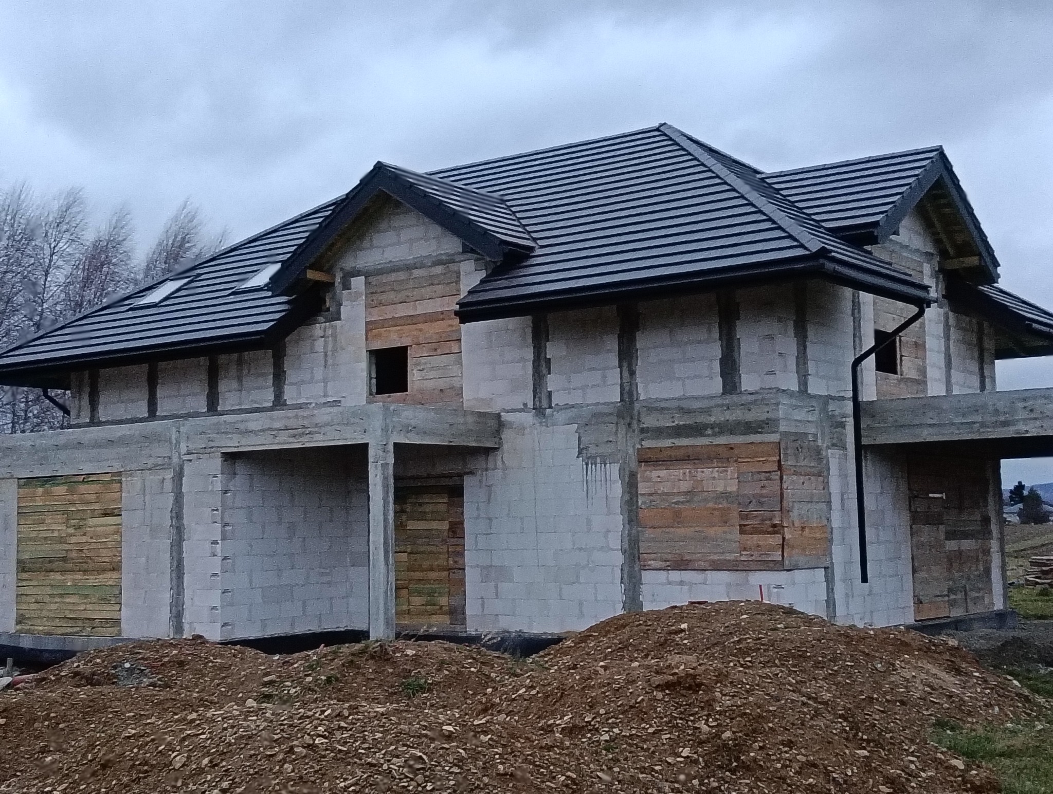 Surowy stan domu jednorodzinnego z szarej kostki, świeżo pokrytego czarną dachówką i orynnowaniem, z deskami zasłaniającymi otwory okienne i drzwiowe, na tle pochmurnego nieba.