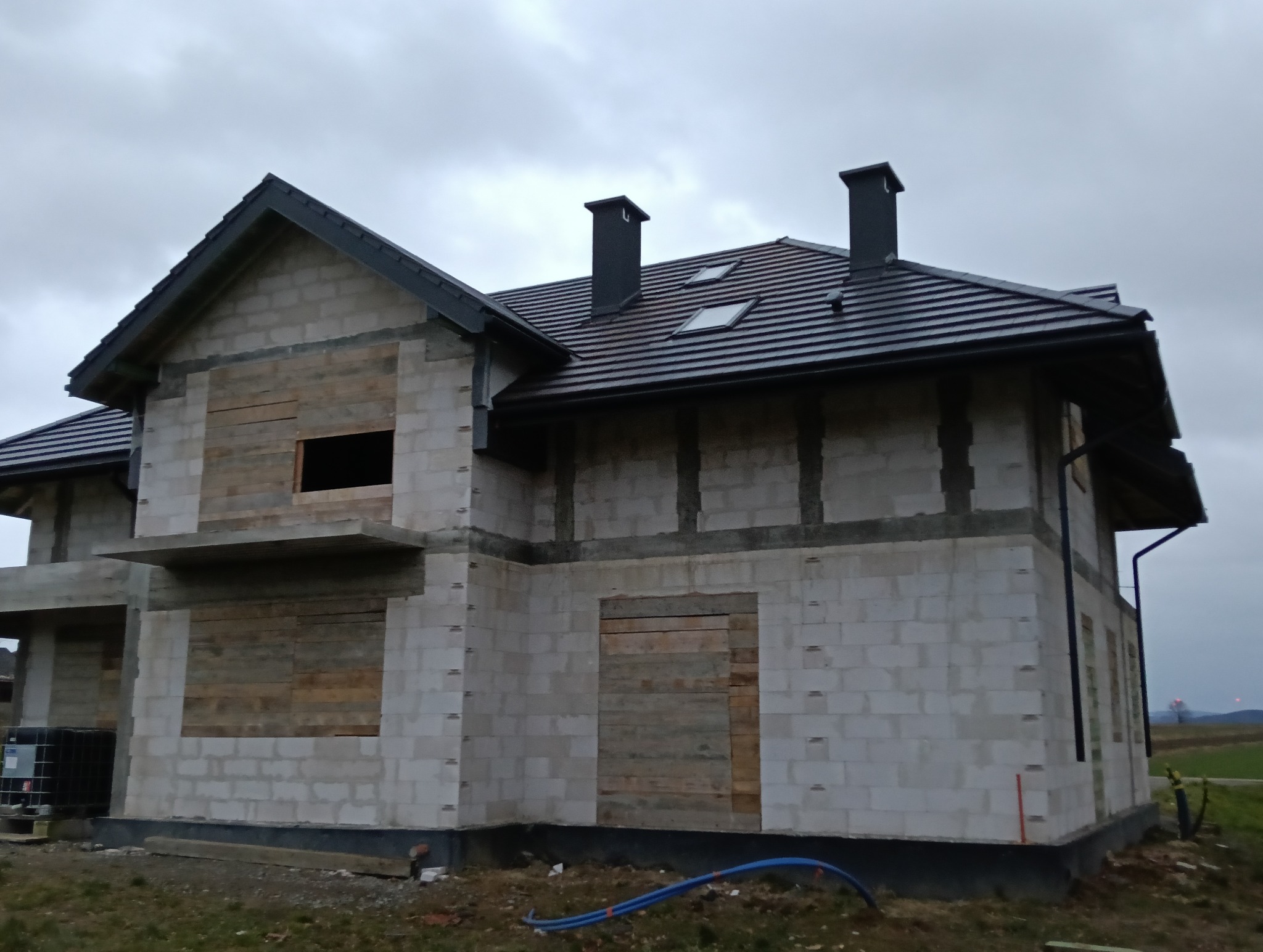 Dom w trakcie budowy, z szarą dachówką, widoczne kominy i okna dachowe, ściany z betonu komórkowego z zabezpieczonymi otworami okiennymi i drzwiowymi deskami.