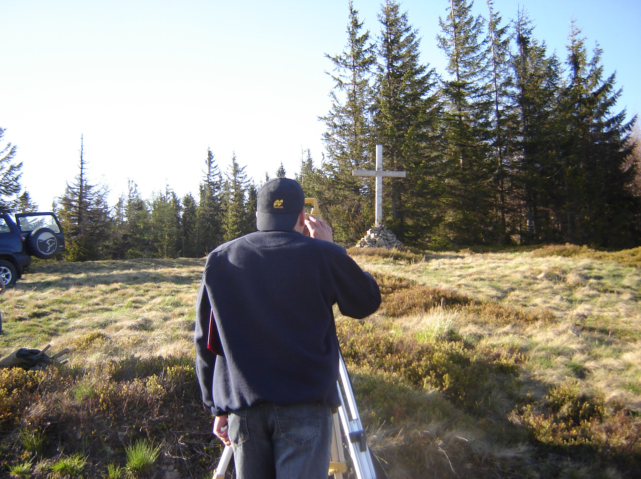 Mężczyzna w bluzie i czapce z daszkiem, stojący tyłem, korzysta z niwelatora na trójnogu na tle górskiego krajobrazu z lasem iglastym i krzyżem na szczycie.