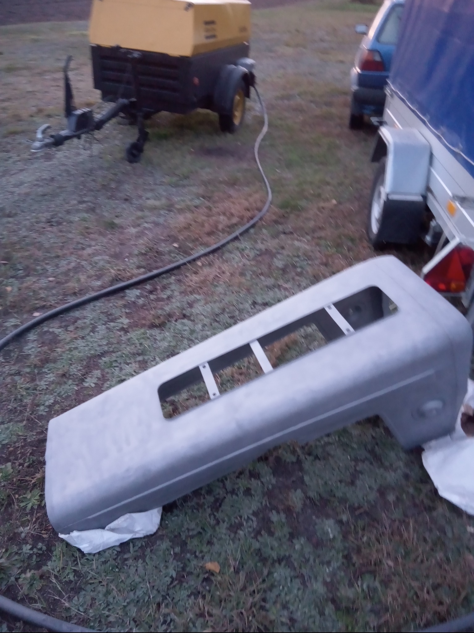 Szary element karoserii samochodu z otworami, prawdopodobnie po piaskowaniu, leżący na trawie obok agregatu prądotwórczego na przyczepce i przyczepy samochodowej.