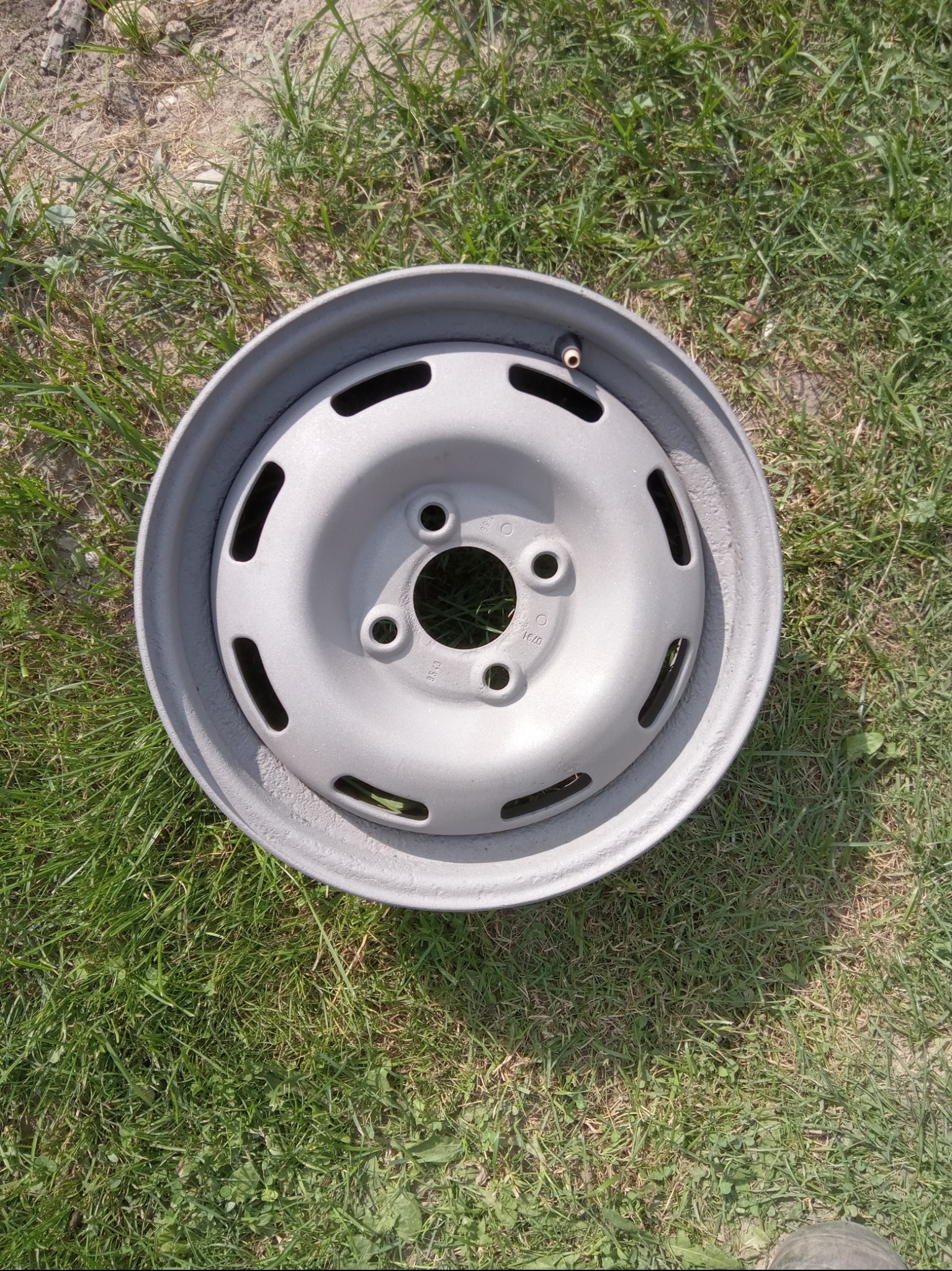 Szara, stalowa felga samochodowa z widocznymi śladami korozji i niedokładnego malowania leży na trawie.
