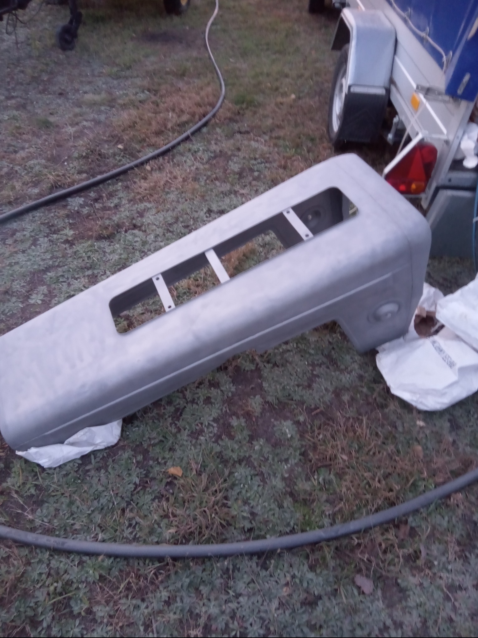 Szary, wstępnie obrobiony element karoserii pojazdu z otworami, leżący na trawie obok przyczepy i worków z materiałem ściernym.