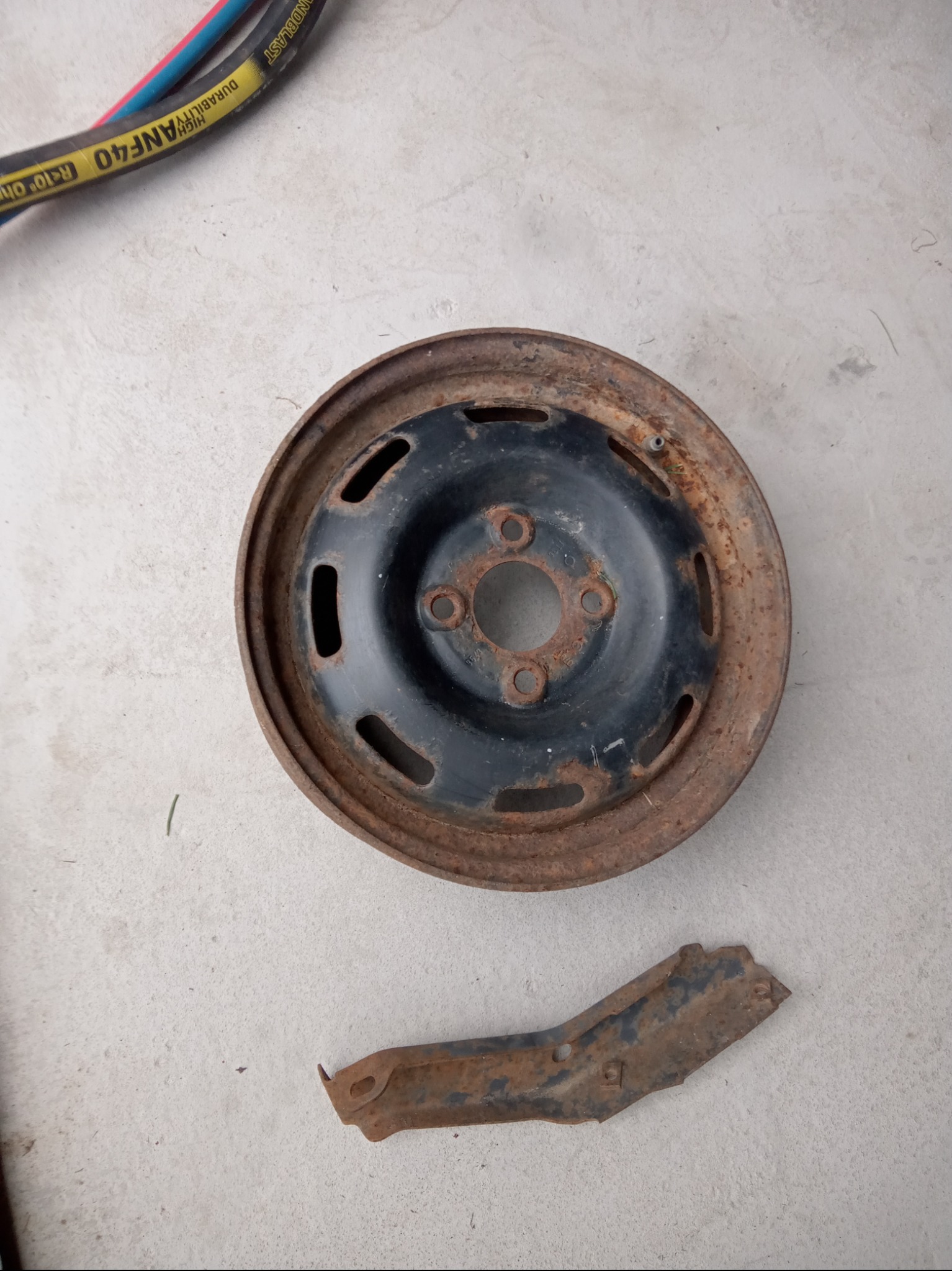 Zardzewiała felga samochodowa z fragmentem skorodowanego elementu metalowego leżące na betonowej posadzce, widoczny wąż do piaskowania w tle.