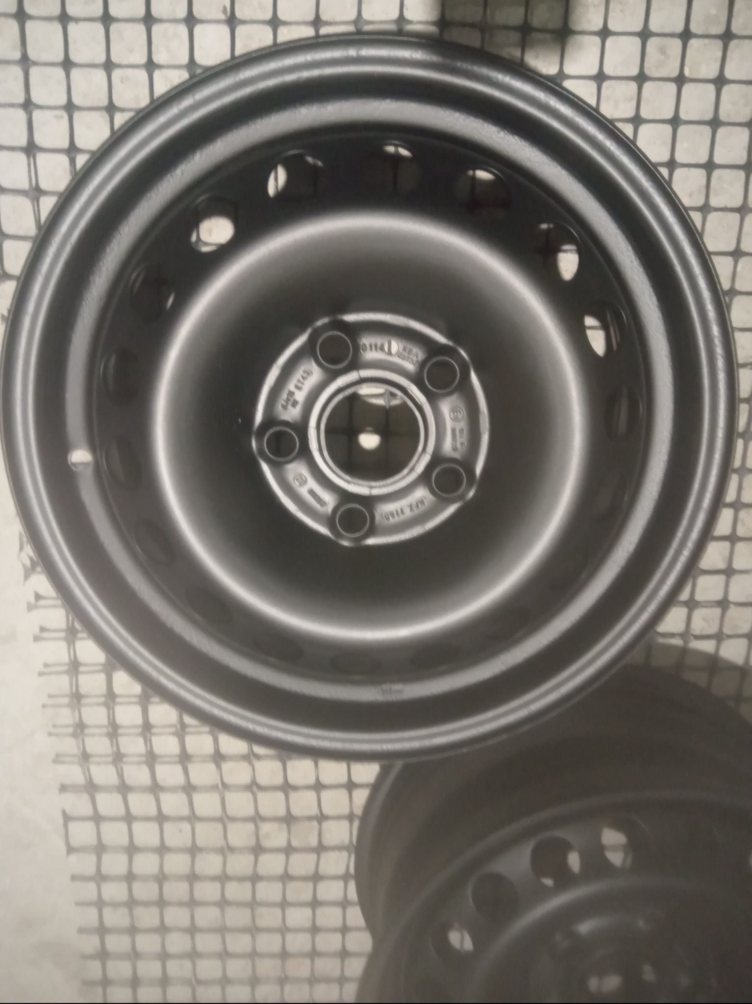 Dwie stalowe felgi samochodowe po piaskowaniu, leżące na siatce, z widocznymi otworami i oznaczeniami producenta.