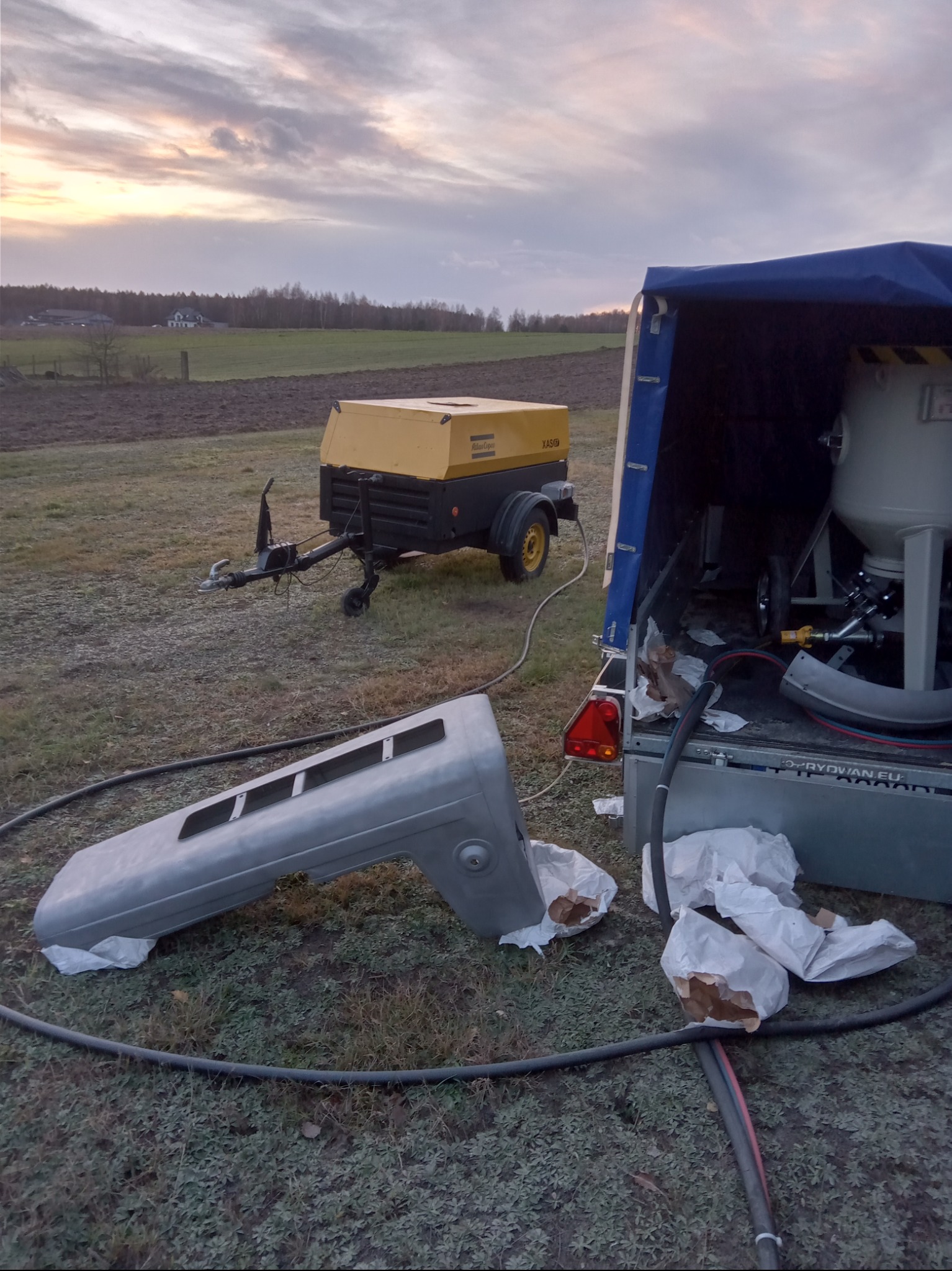 Przygotowany do piaskowania element karoserii samochodu leży na trawie obok przyczepy z kompresorem i piaskarką w plenerze o zmierzchu.