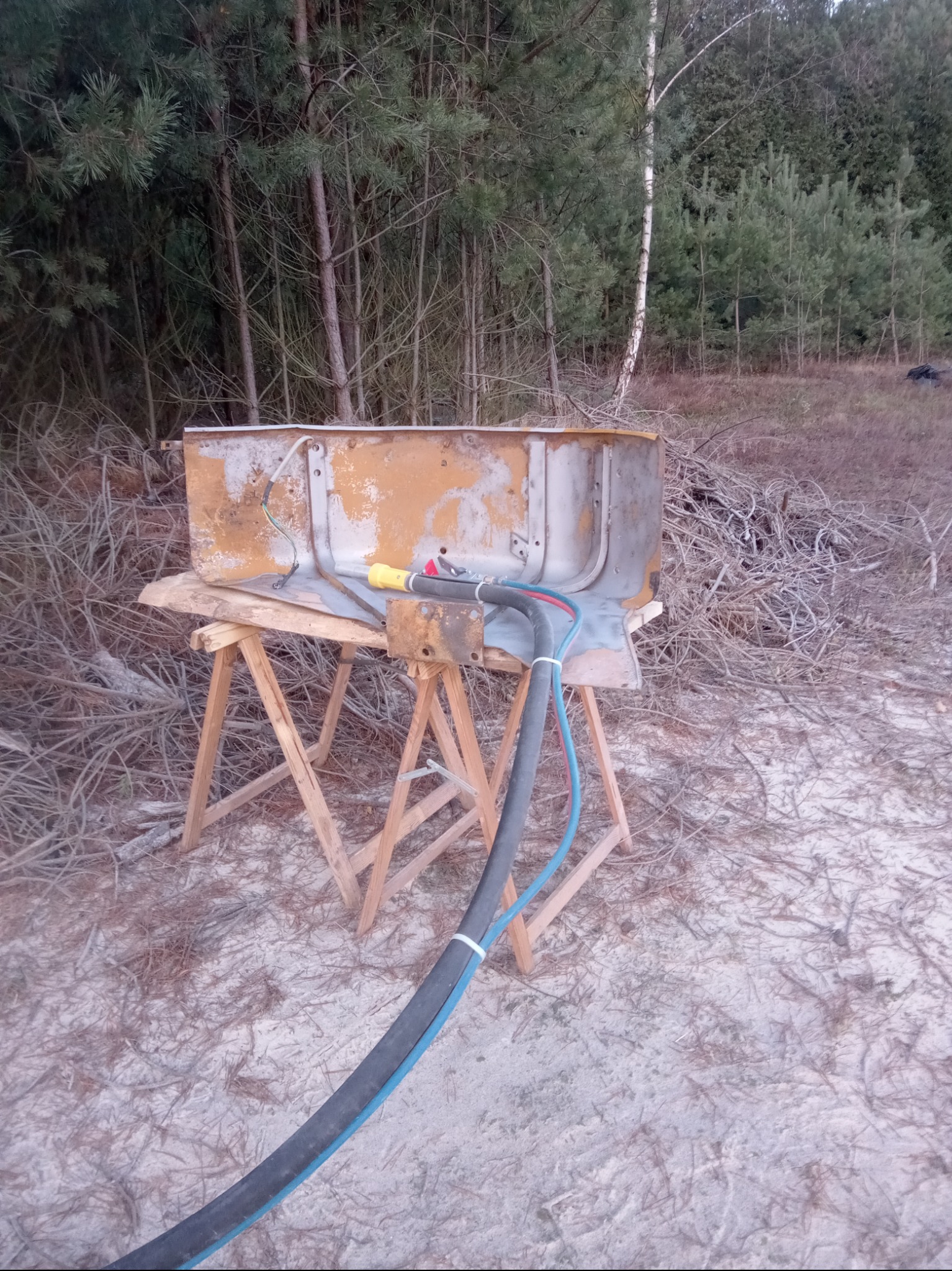 Metalowa wanna po obróbce, częściowo oczyszczona z rdzy, umieszczona na drewnianych kozłach, z podłączonym przewodem do piaskowania, w tle las i gałęzie.