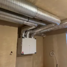 Centrala wentylacyjna z rekuperacją zamontowana na ścianie, z widocznymi przewodami wentylacyjnymi w aluminiowej izolacji, surowy stan wykończenia wnętrza.