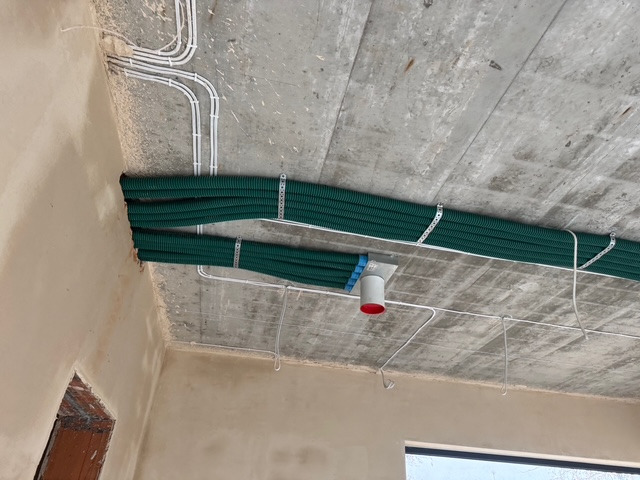 Zielone rury wentylacyjne przymocowane do surowego betonowego sufitu za pomocą metalowych uchwytów, widoczne również białe rurki instalacyjne i fragment otworu wentylacyjnego z czerwoną wstawką.