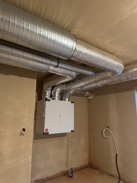 Centrala rekuperacyjna z podłączonymi przewodami wentylacyjnymi w surowym pomieszczeniu z widocznymi niedoskonałościami na ścianach i podłodze.