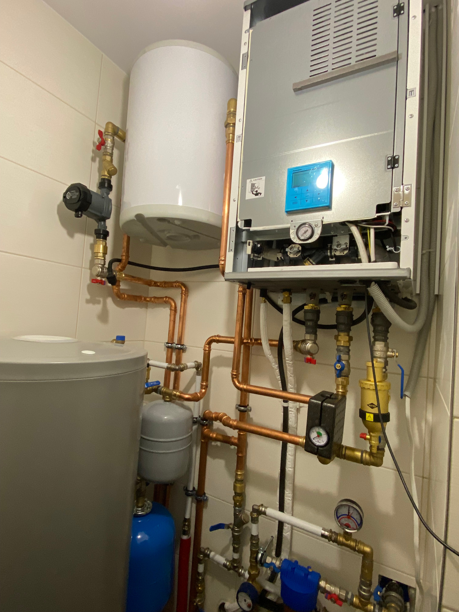 Złożona instalacja grzewcza w kotłowni: zbiornik ciepłej wody użytkowej, pompa obiegowa, naczynia wzbiorcze i piec gazowy kondensacyjny z otwartą obudową, widoczne liczne rury miedziane i zawory.