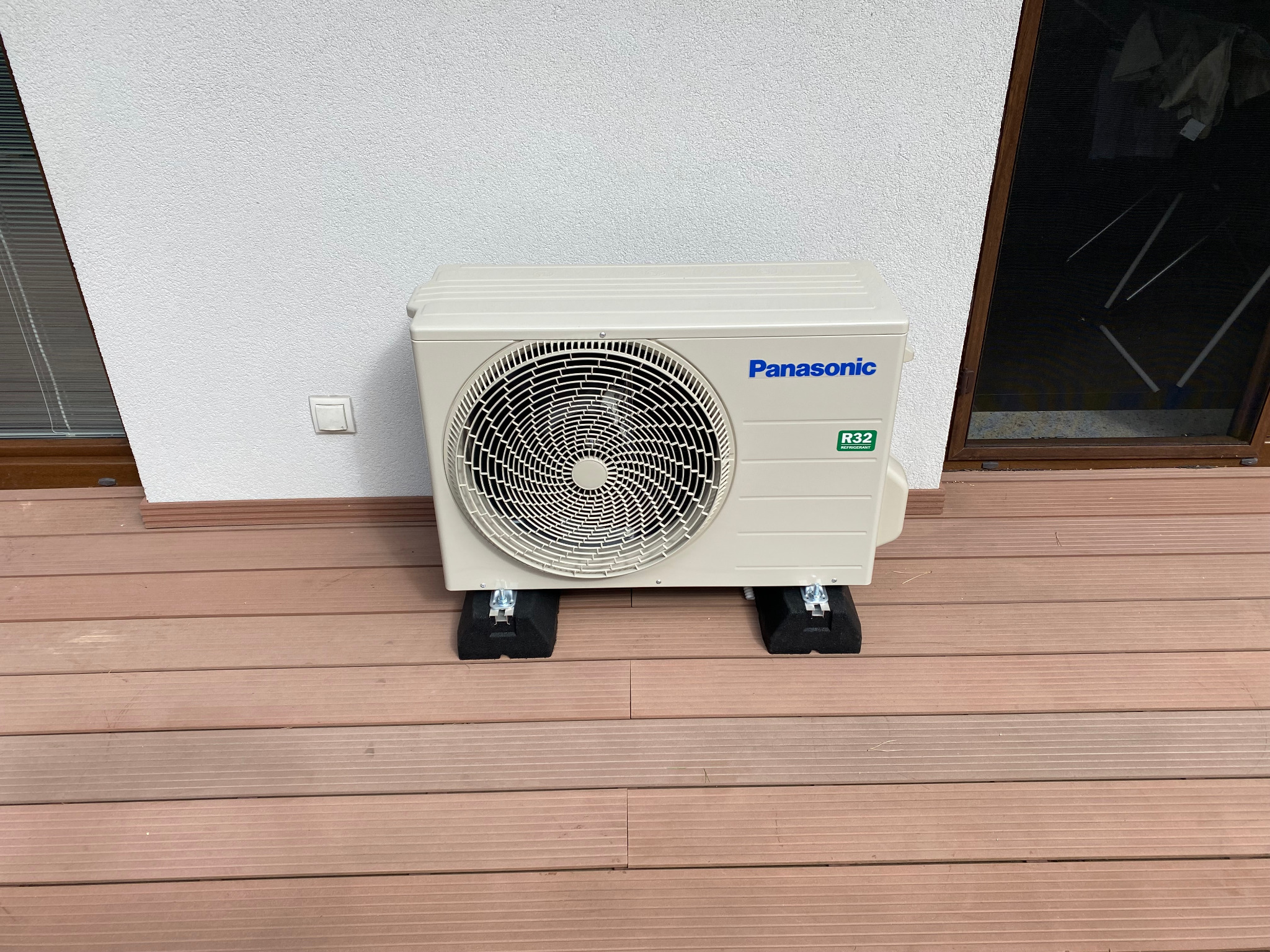 Zewnętrzna jednostka klimatyzatora Panasonic R32 na drewnianym tarasie, zamontowana na czarnych podkładkach antywibracyjnych, biała ściana z kontaktem i brązowa rama okienna w tle.