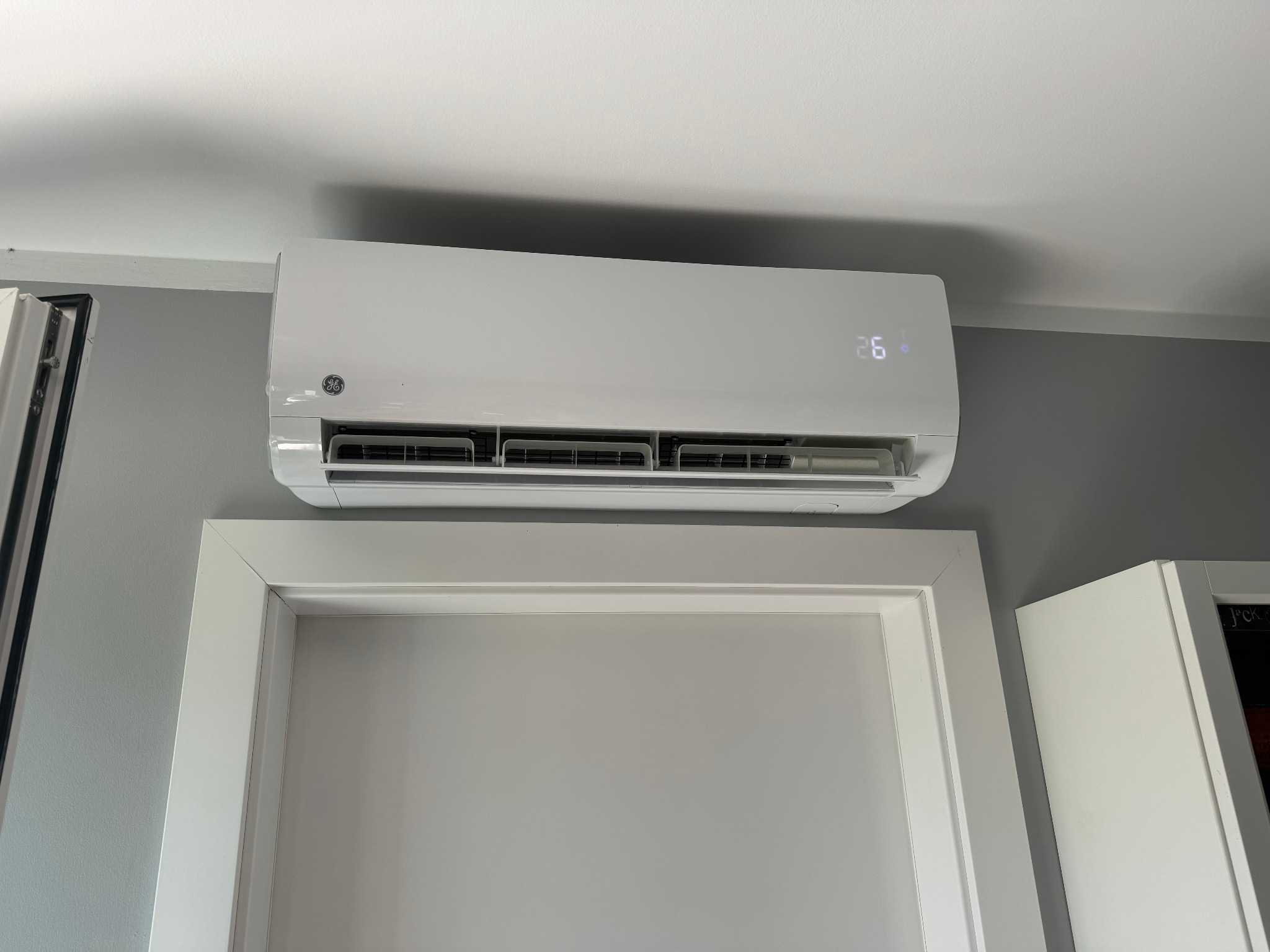Biała jednostka klimatyzacji typu split zamontowana nad drzwiami w szarym pomieszczeniu, wyświetlająca temperaturę 26 stopni Celsjusza.