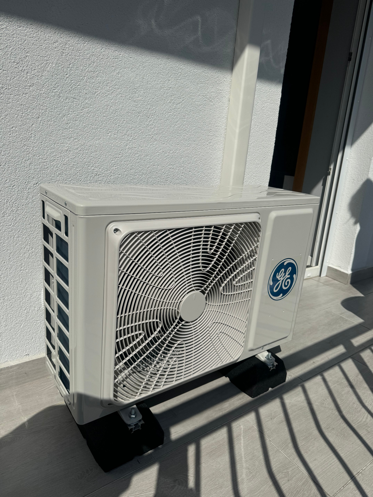 Zewnętrzna jednostka klimatyzatora marki GE, zamontowana na balkonie z widocznym cieniem balustrady, umieszczona na czarnych podkładkach antywibracyjnych.