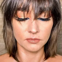 Portret kobiety z brązowymi włosami i wyrazistym makijażem oczu z niebieską kreską, patrzącej w dół, na neutralnym tle.