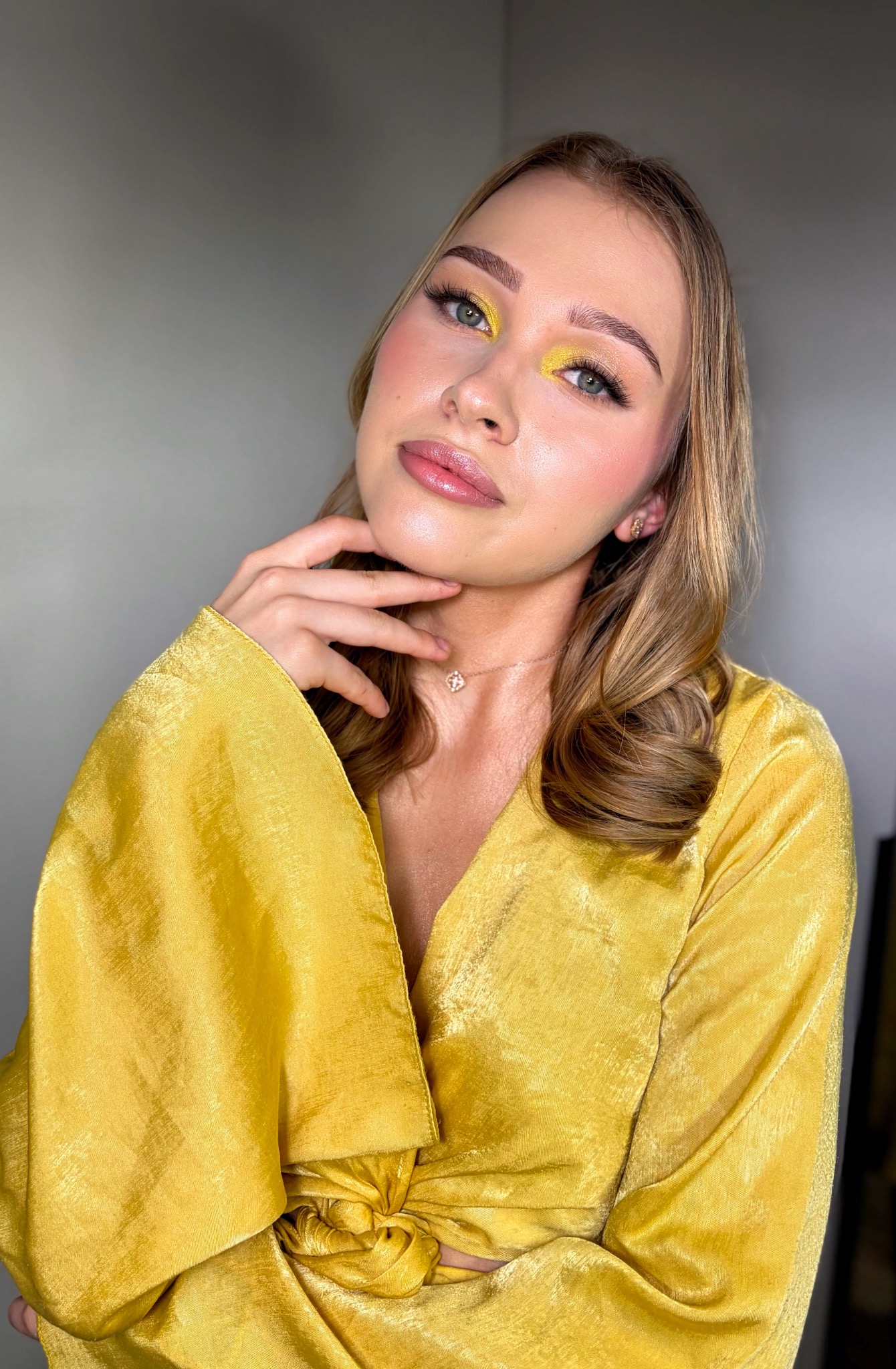 Portret kobiety z żółtym cieniem na powiekach i błyszczącymi ustami, w złotej sukience, pozującej z dłonią pod brodą na tle szarej ściany.