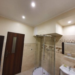 Piotr Marcinkowski - Wykończona łazienka w beżowych odcieniach z kabiną prysznicową, umywalką z szafką i lustrem z dekoracyjnymi elementami. Widoczna drewniana ościeżnica drzwiowa i fragment niezamontowanego skrzydła...