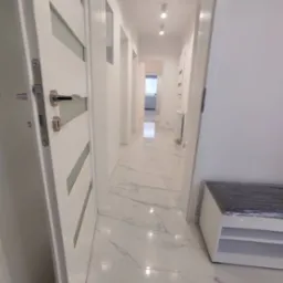 Biały korytarz z marmurową podłogą, widok na wnętrze przez otwarte drzwi, minimalistyczny styl, z widoczną ławką z tworzywa.