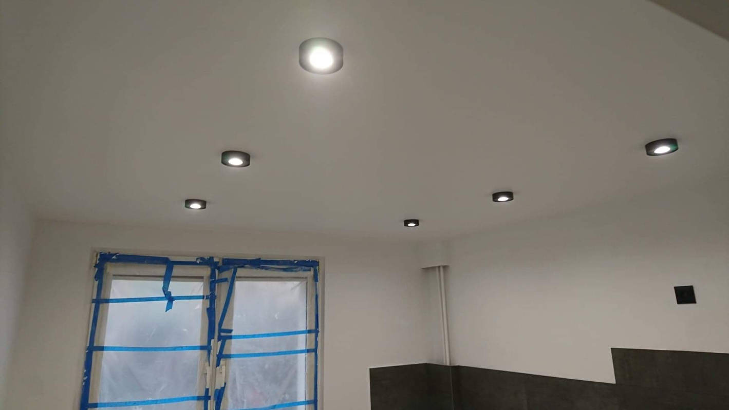 Biały sufit z zamontowanymi sześcioma okrągłymi lampami LED, okno zabezpieczone folią malarską i taśmą.