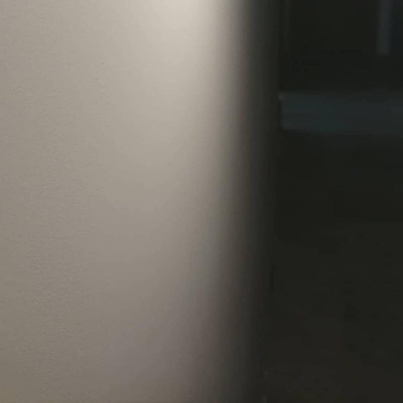 Świeżo pomalowana ściana w odcieniach szarości z wyraźnym podziałem na oświetloną i zacienioną część, widoczny fragment ciemnego pomieszczenia w tle.