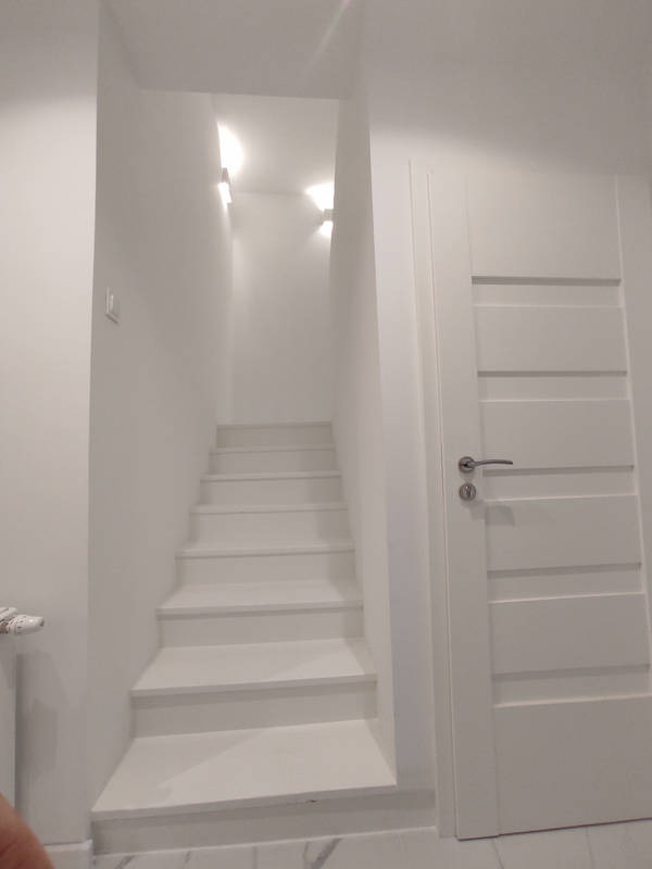Białe schody prowadzące do góry, oświetlone kinkietami, świeżo po odnowieniu w domu w Żarach, widoczne z bliska, obok białe drzwi z poziomymi panelami.