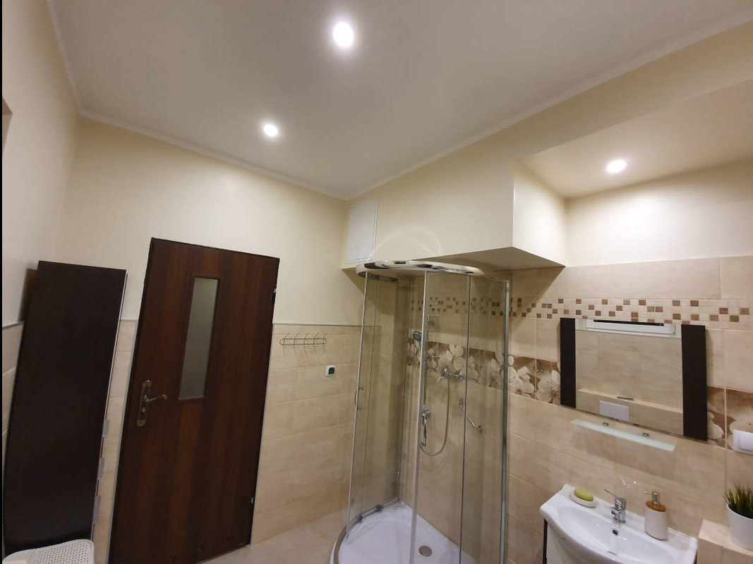 Wykończona łazienka w beżowych odcieniach z kabiną prysznicową, umywalką z szafką i lustrem z dekoracyjnymi elementami. Widoczna drewniana ościeżnica drzwiowa i fragment niezamontowanego skrzydła...