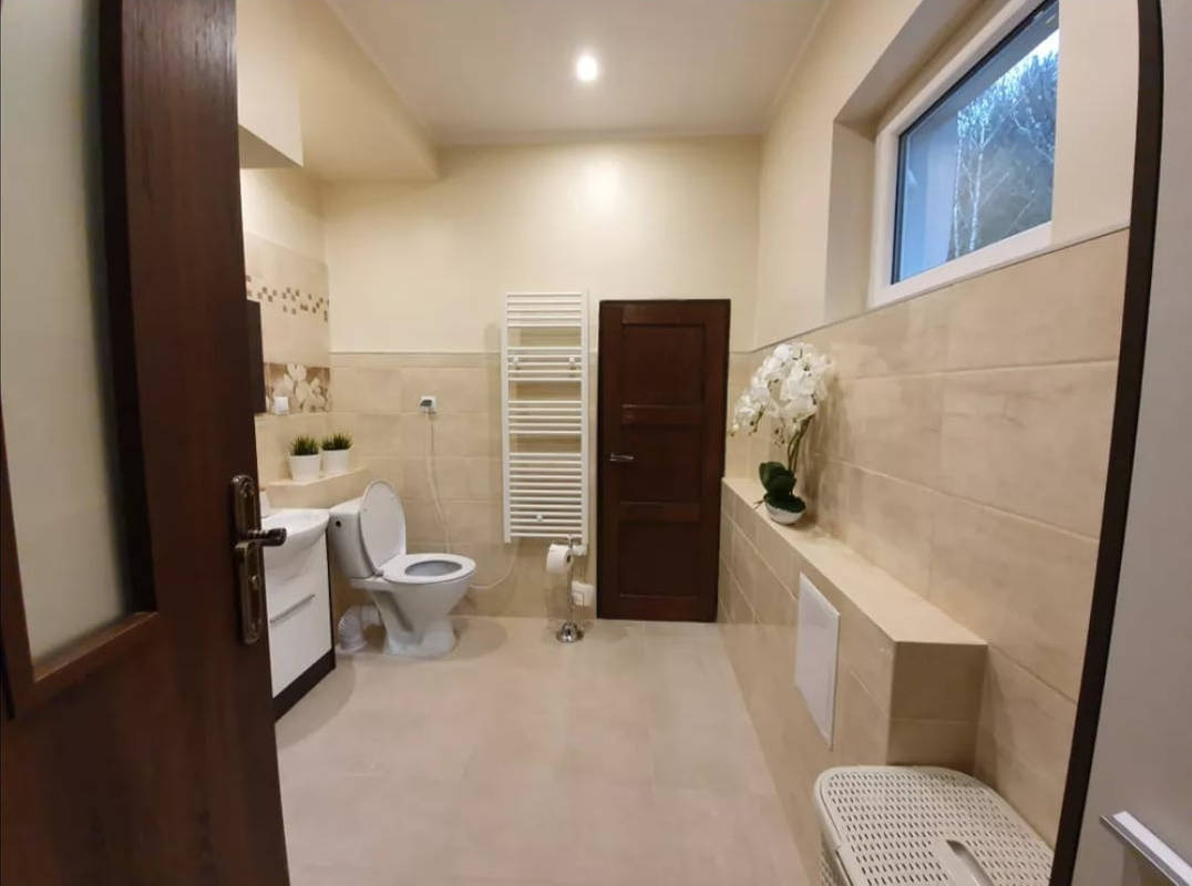 Jasna łazienka z beżowymi płytkami, białą ceramiką i grzejnikiem drabinkowym, widok zza uchylonych, ciemnych drzwi.