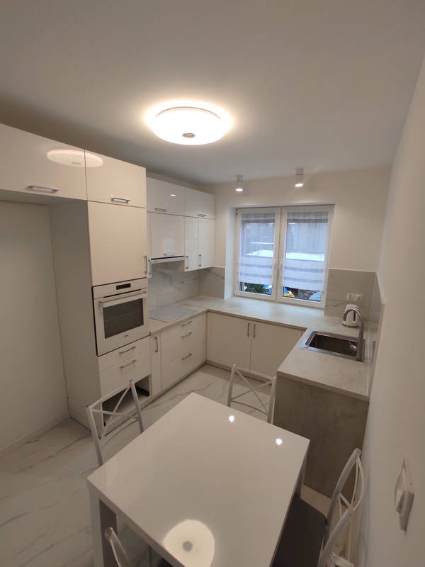 Jasna, nowocześnie urządzona kuchnia z białymi błyszczącymi szafkami, marmurową podłogą i stołem z krzesłami. Okno z roletami częściowo zasłonięte. Widoczny fragment blatu z szarym wykończeniem.