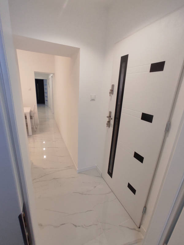 Wykończony korytarz z białą podłogą imitującą marmur, białe ściany i drzwi z czarnymi akcentami, widok w głąb mieszkania z białymi meblami w oddali.