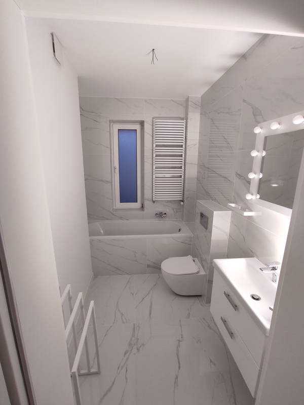 Wnętrze wykończonej łazienki w bieli i szarości z marmurowymi płytkami na podłodze i ścianach; widoczna wanna, toaleta, umywalka z szafką oraz grzejnik drabinkowy. Na suficie widoczne niedokończone...